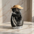 Hugging Couple Figurine - A sculptural decorative figurine of two embracing figures with a woven straw hat, shown on a light marble surface against a marble wall.
