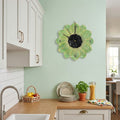Flower Shaped Clock - A green and yellow sunflower-shaped wall clock with a black centre is mounted on a mint green kitchen wall above a wooden countertop with plates, a basket, and a potted plant.