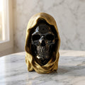 Hooded Skull Figurines - A decorative skull sculpture with a glossy black face and a draped metallic gold hood, displayed on a white marble surface against a light marble wall.