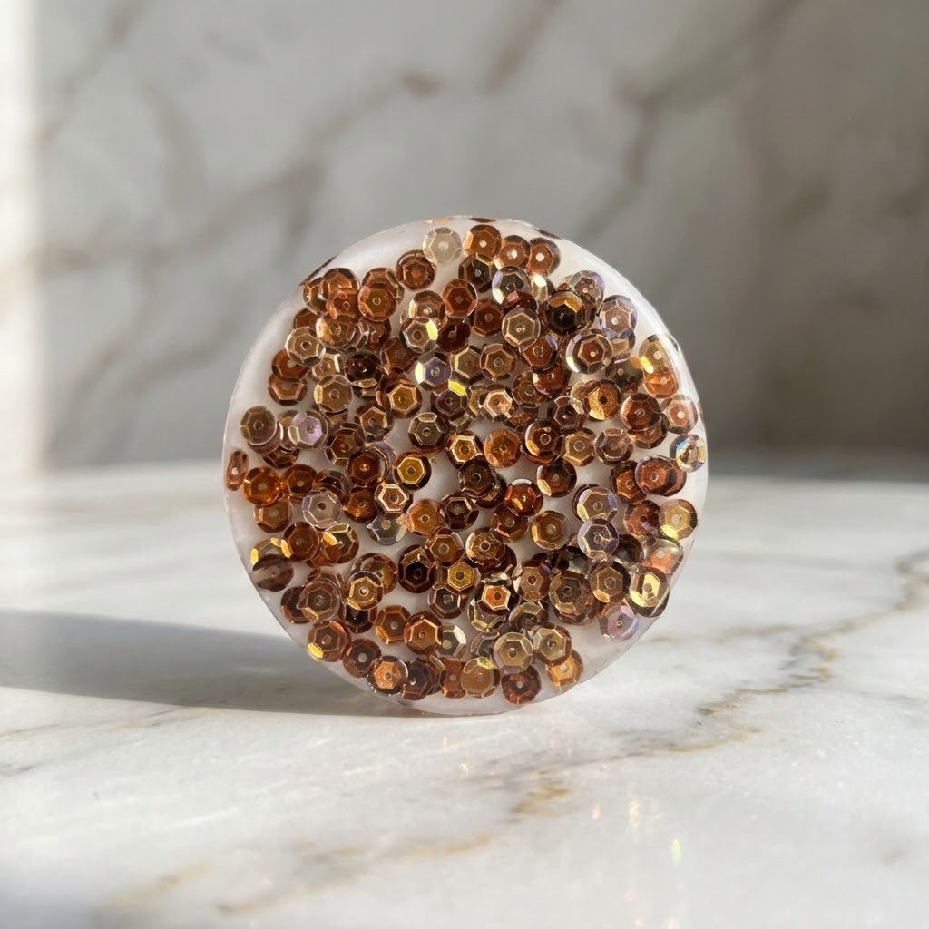 Golden Sequin Coasters - A round resin coaster filled with small amber-brown hexagonal sequins, photographed on a light marble surface with a marble background.