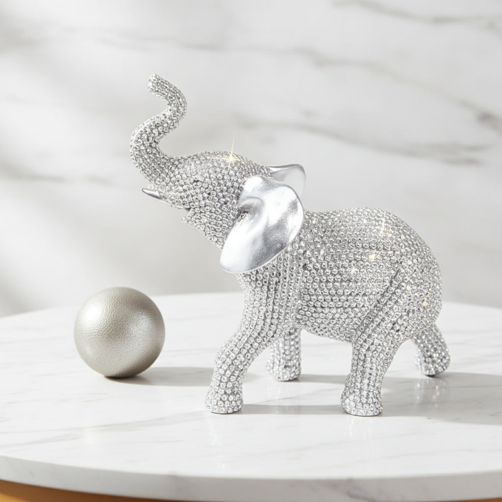 A rhinestone-covered elephant figurine in silver stands on a white marble surface beside a matching silver sphere, with a soft marble background.