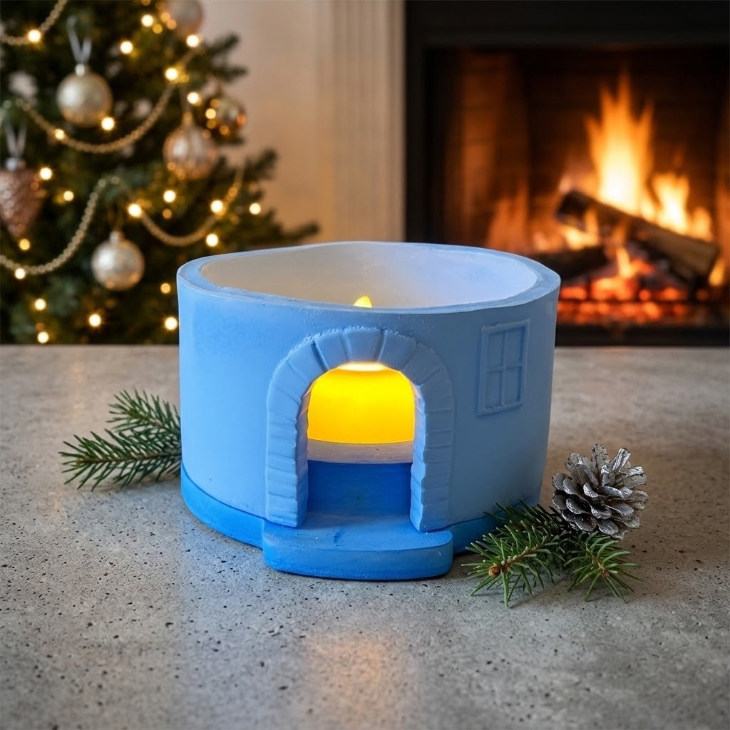 Candle Holder Houses - A small blue house-shaped candle holder with a glowing yellow tea light inside, styled on a tabletop in front of a fireplace with a Christmas tree in the background.