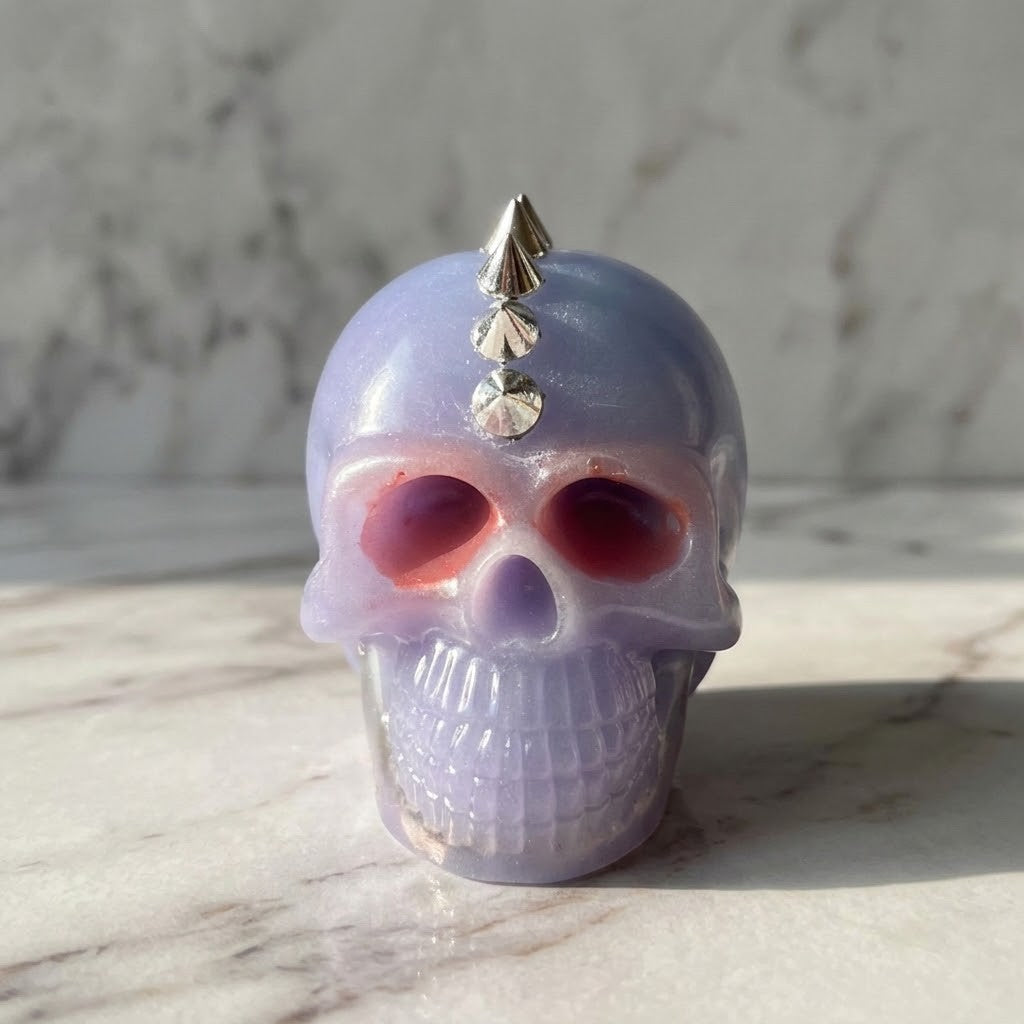 Studded Gothic Skull - A small glossy skull-shaped object in pale lavender with metallic silver spikes on top, photographed on a light marble surface against a marble background.