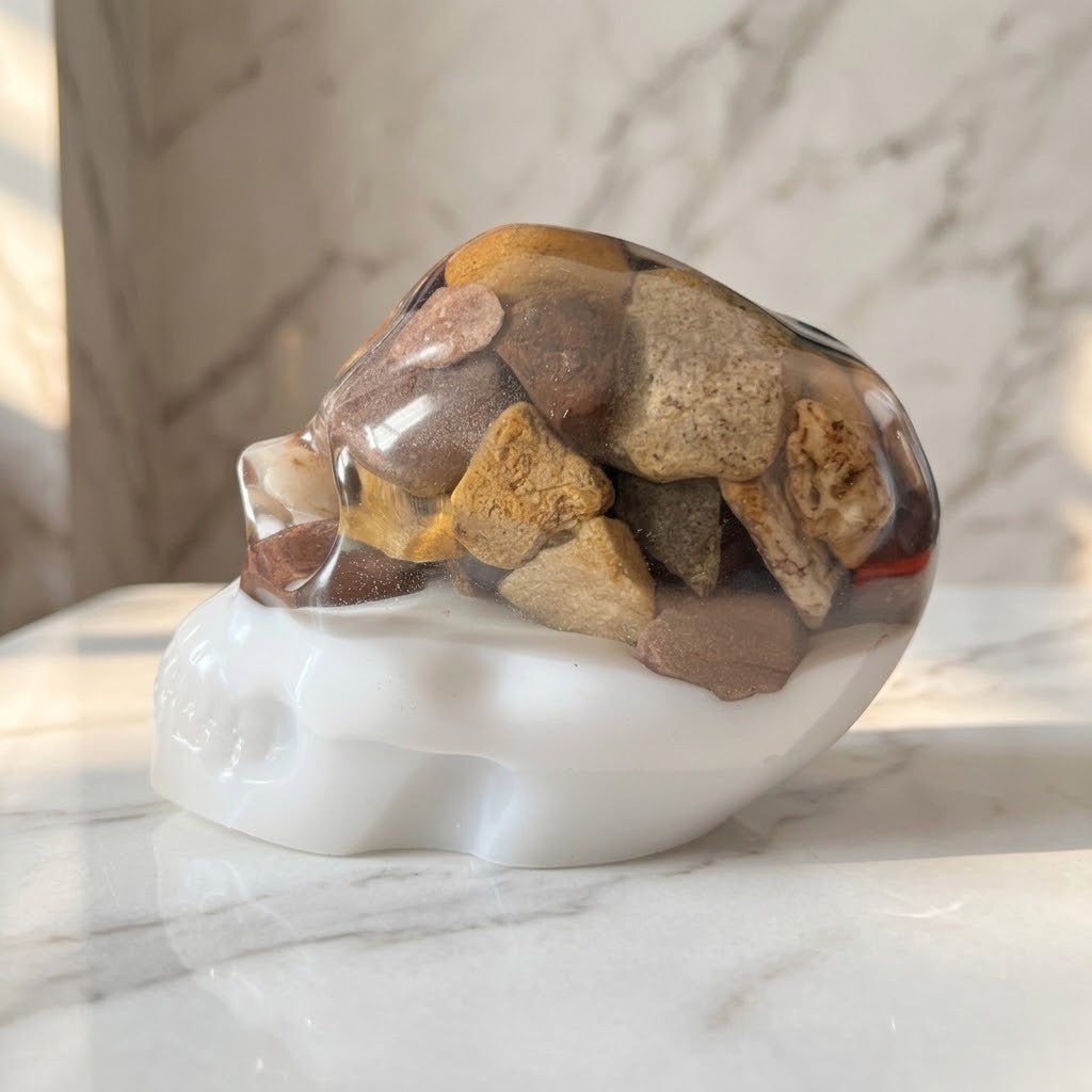 Gothic Resin Stone Skull - Side view of a polished resin skull figurine resting on a white marble surface. The skull features a glossy white base and a transparent upper section filled with smooth river stones in earthy brown, tan, and grey tones.